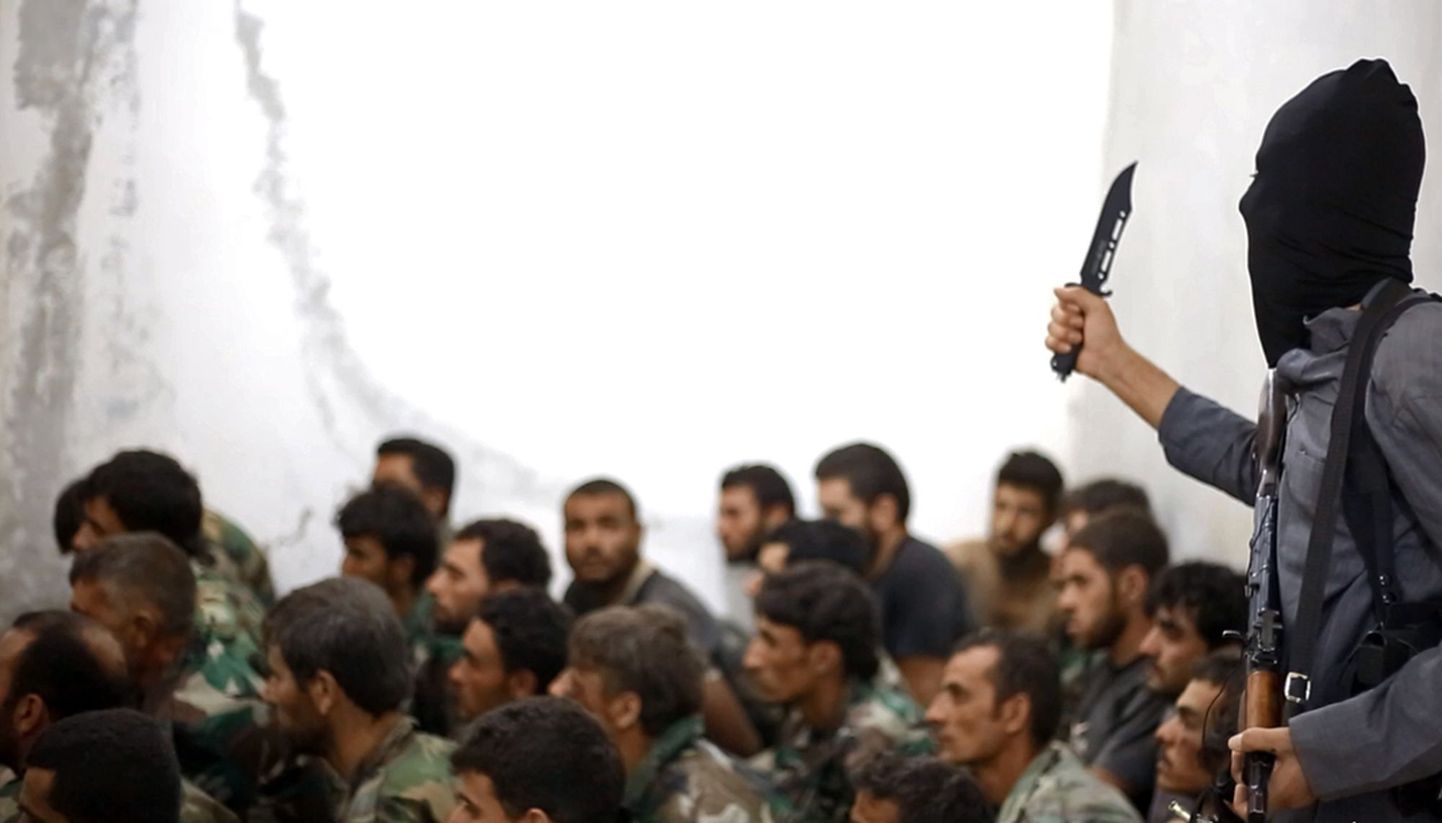 На снимке, распространенном пресс-центром Исламского государства, запечатлен боевик ИГ на фоне захваченных в плен сирийских солдат.