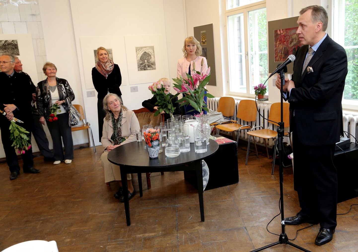 Kaks kunstnikku: poeg Aapo Pukk on mikrofoni juures ja ema Laine Pukk kuulab teda laua taga, mis on kaetud lillede ootel vaasidega.