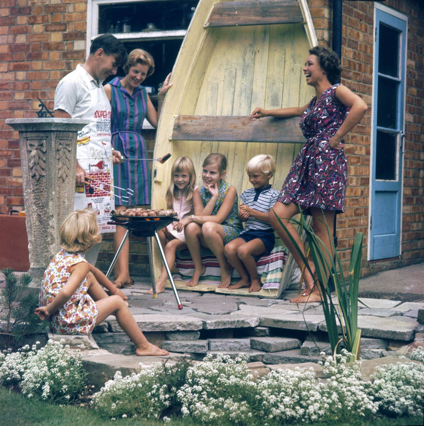 A happy family scene as Dad  barbecues the food on a tripod  barbeque on the patio and  three of the children use an  upturned rowing boat as a  seat!     Date: 1969