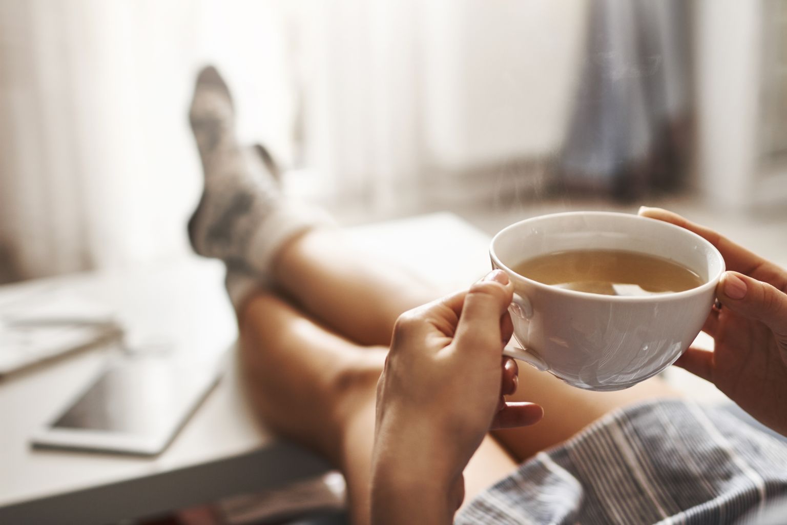 Cup of tea and chill. Woman lying on couch, holding legs on coffee table, drinking hot coffee and enjoying morning, being in dreamy and relaxed mood. Girl in oversized shirt takes break at home
