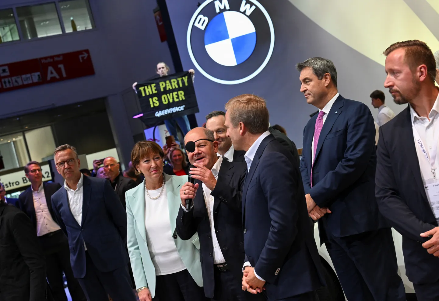 Saksamaa liidukantsler Olaf Scholz Müncheni autonäitusel 2023. aastal. Taustal seisab plakatiga Greenpeace'i protestija.