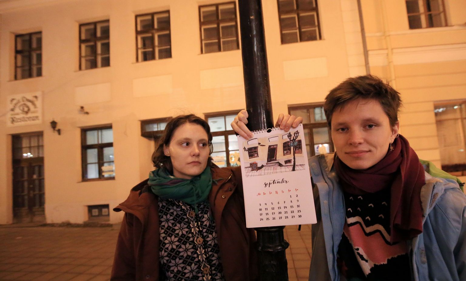 Maša Kanatova (vasakult) ja Saša Miljakina seisavad Tartu raeplatsi serval, selja taga saiapoe aknad ja uks ning käes nende endi valmistatud kunstikalender aastaks 2018 just sellelt leheküljelt, kus on saiapoe joonistus.