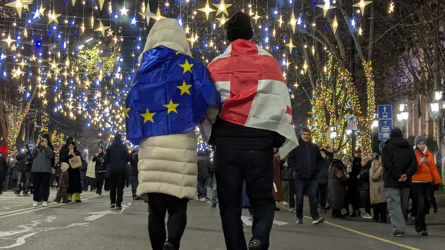Два человека сфотографированы со спины, накрылись флагами Грузии и ЕС и идут по улице под гирляндами в виде звезд