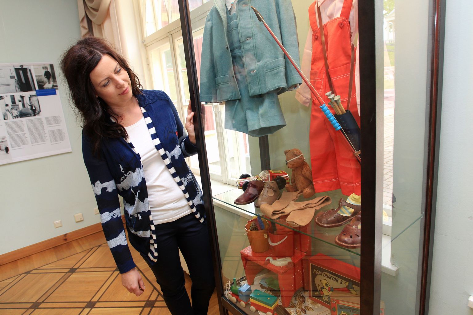 Näituse kuraator Kristiina Tael vitriini ees, kuhu on välja pandud kunagised riided ja mänguasjad.