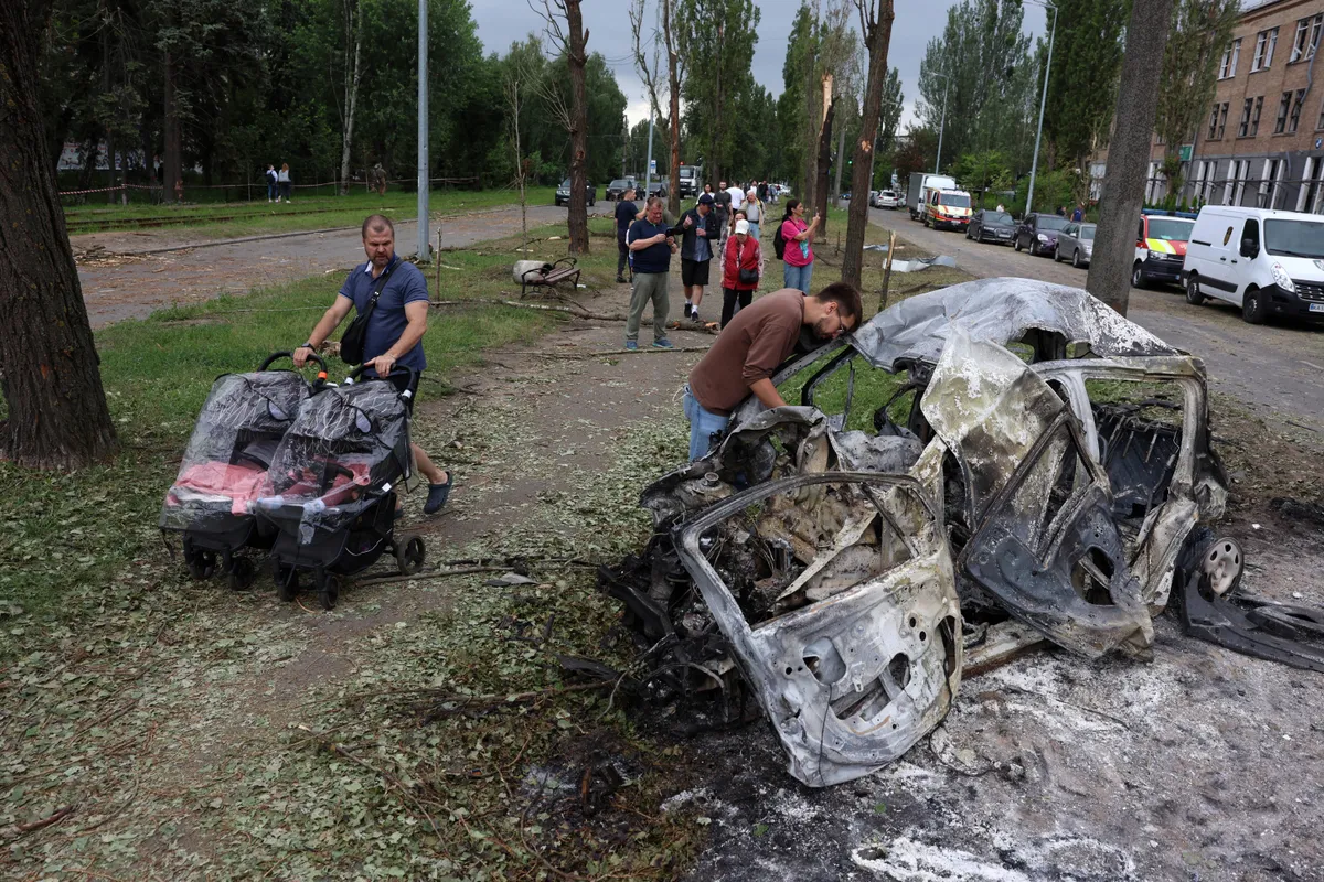 A man pushes two baby strollers past a car burned during a Russian missile attack overnight in Kyiv, June 17, 2025. Ukrainian authorities continue conducting rescue operations following a ballistic missile and drone bombardment which killed 14 people and wounded over 50.