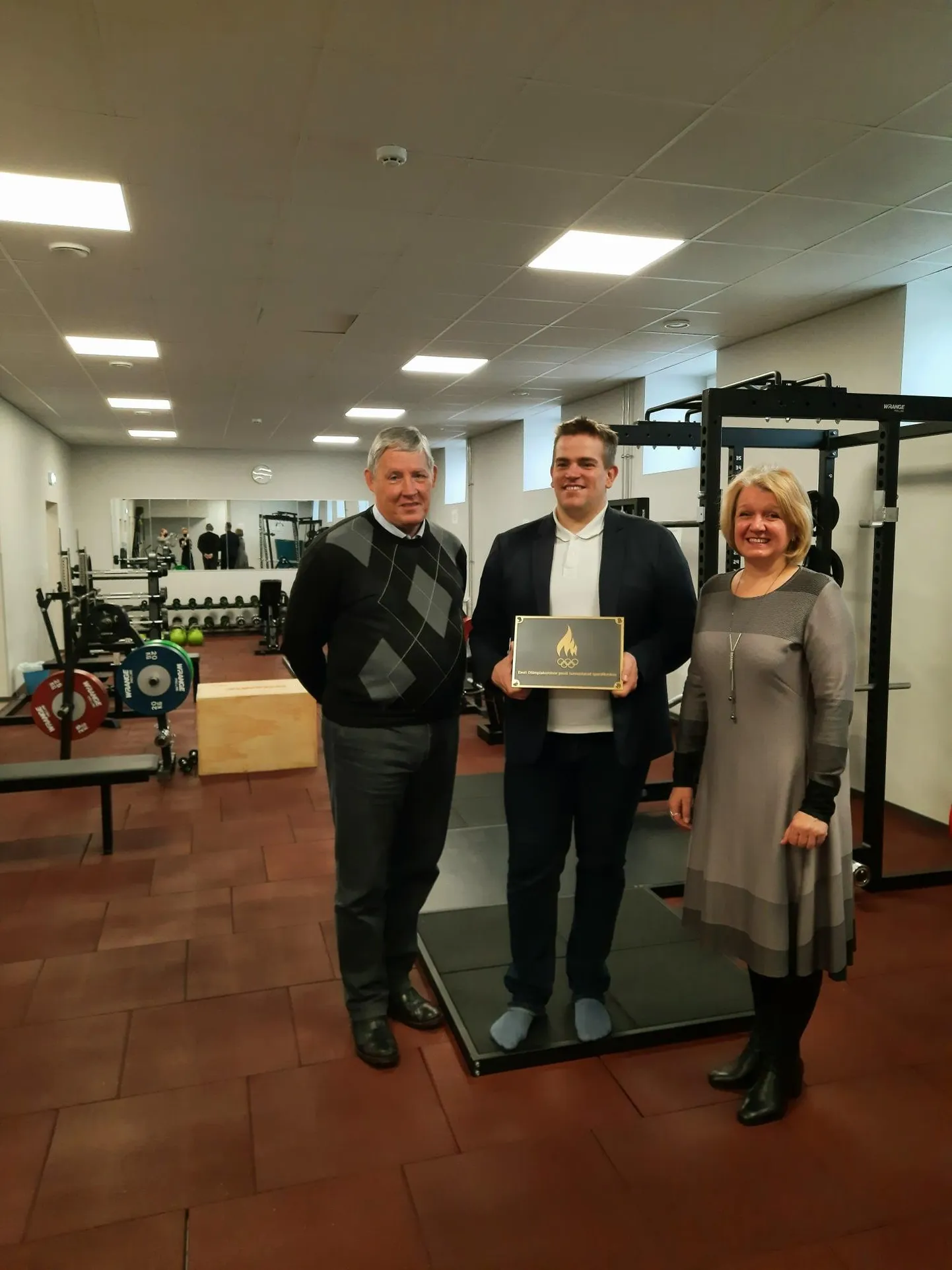 Lääne-Virumaa Spordiliidu juhatuse liige Erich Petrovits (vasakult), Kadrina spordikeskuse juhatuse liige Robert Salep ja Kadrina vallavanem Kairit Pihlak rõõmustavad jõusaalis vastse autasu üle.